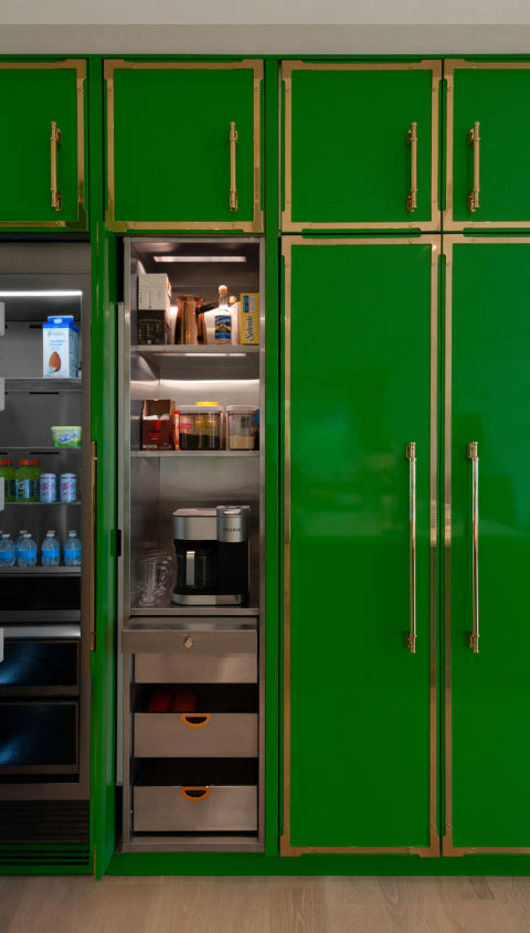 Close-up of built-in green kitchen cabinets with gold trim, showing a section with an open refrigerator and an illuminated pantry with a built-in coffee maker.