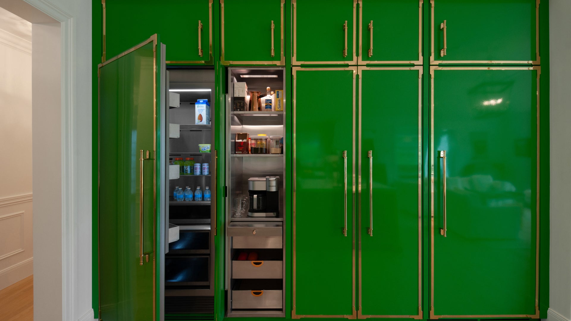 A full wall of built-in kitchen cabinetry in a bold green with gold trim and hardware. The open sections reveal a refrigerator with drinks and a coffee station with drawers.