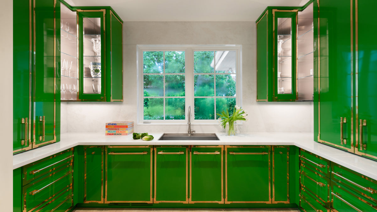 Modern kitchen with striking glossy green cabinets and gold trim. A clean white countertop and backsplash surround a sink under a large window.