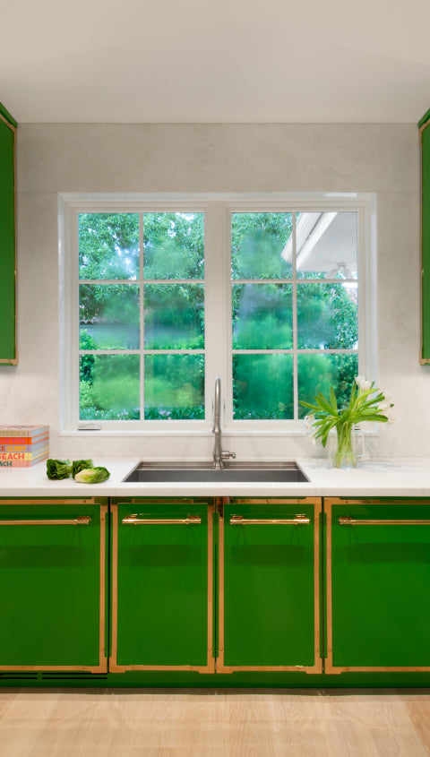 Vibrant kitchen sink area with glossy green cabinets, gold trim, and matching hardware. A white marble countertop surrounds a stainless steel sink, centered under a large window with a view of a lush green outdoor space.
