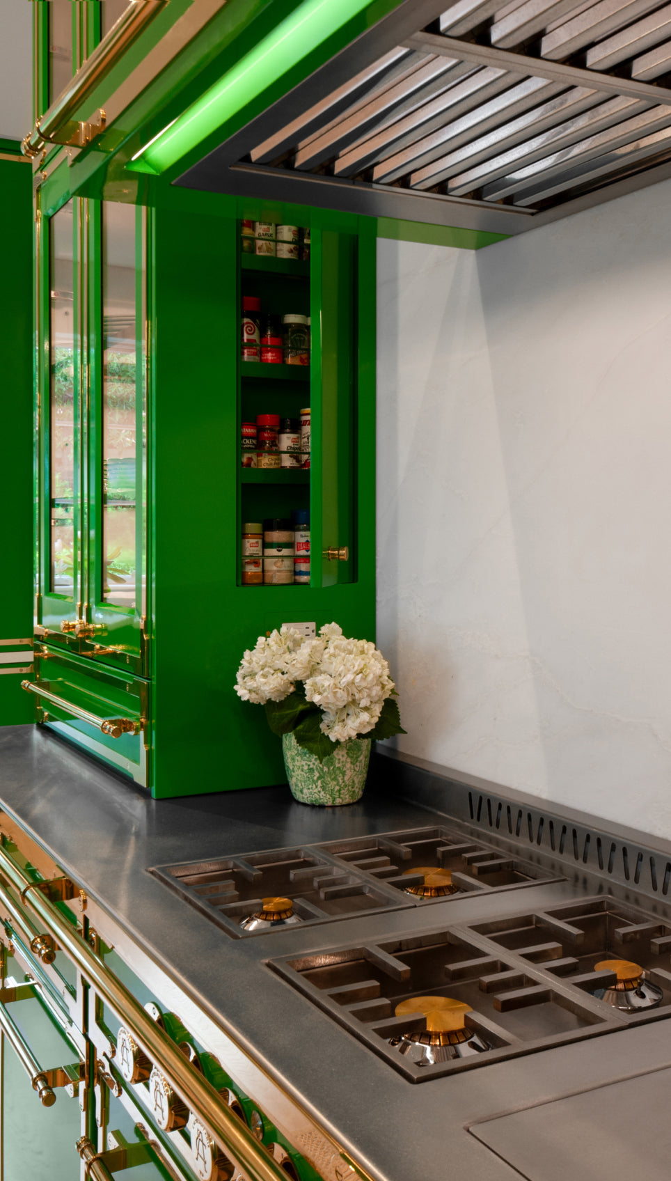 L'Atelier Paris vibrant emerald green custom kitchen cabinetry with polished brass trim, featuring a built-in spice organizer, professional stainless steel cooktop, and matching range hood.