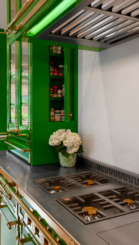 Detailed shot of a modern kitchen with bold green cabinets and gold trim. The image highlights a stainless steel stovetop and an open spice cabinet filled with jars, with a vase of white flowers on the adjacent counter.