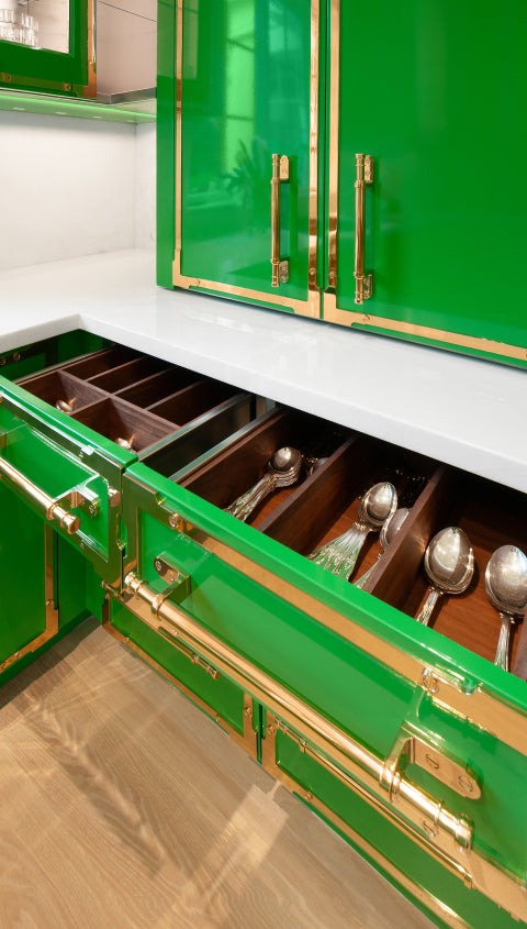A detail shot of a custom kitchen drawer with a glossy green finish and gold hardware. The drawer is open, revealing a walnut wood cutlery organizer with polished silverware.