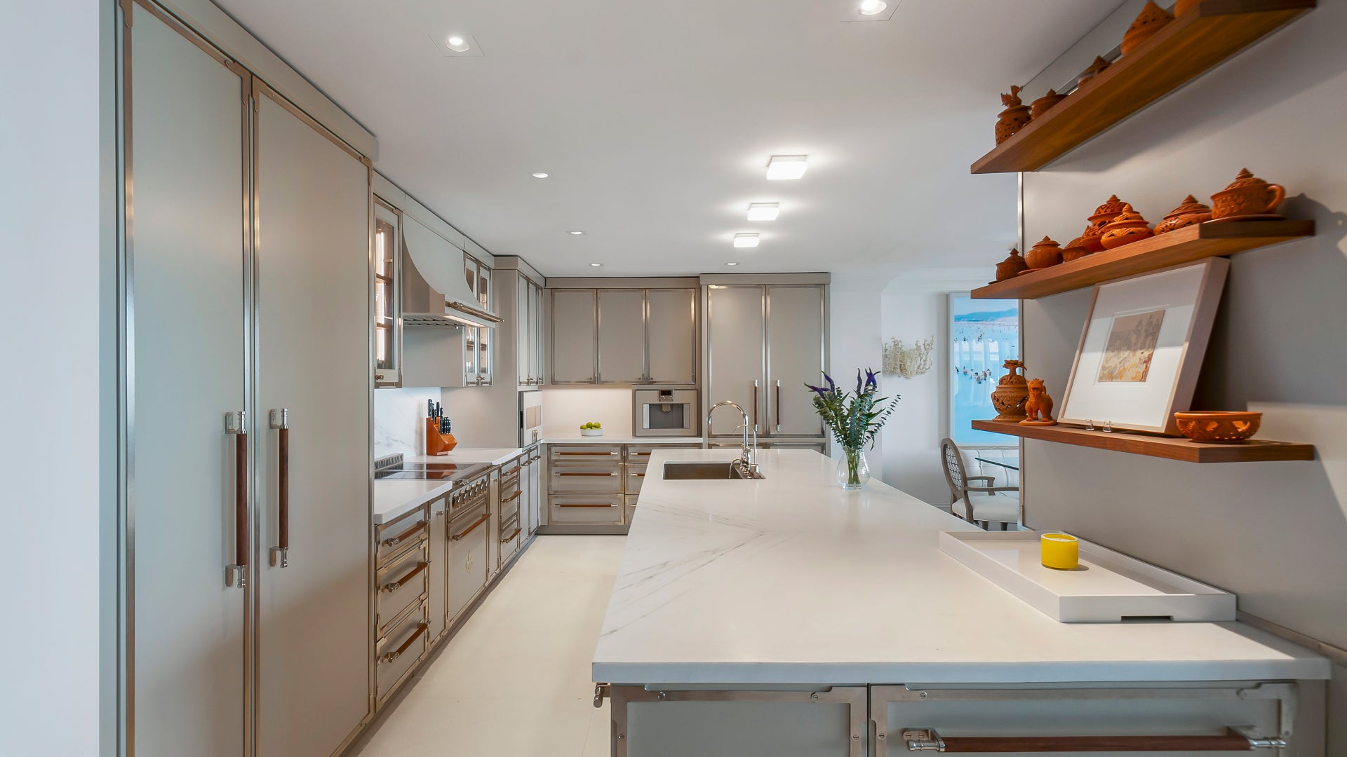 Luxury galley kitchen with polished light gray metallic cabinets, white marble-look quartz countertops, modern stainless steel and brass appliances, and an open dining area