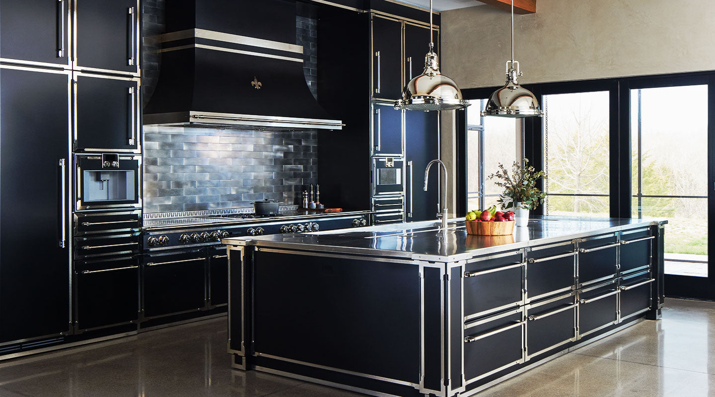 Ultra-modern luxury kitchen featuring all-black cabinetry and a large kitchen island with sleek stainless steel or chrome trim and hardware, a black La Cornue range and custom black range hood with a Fleur de Lis accent, an industrial metal subway tile backsplash, and two chrome pendant lights.