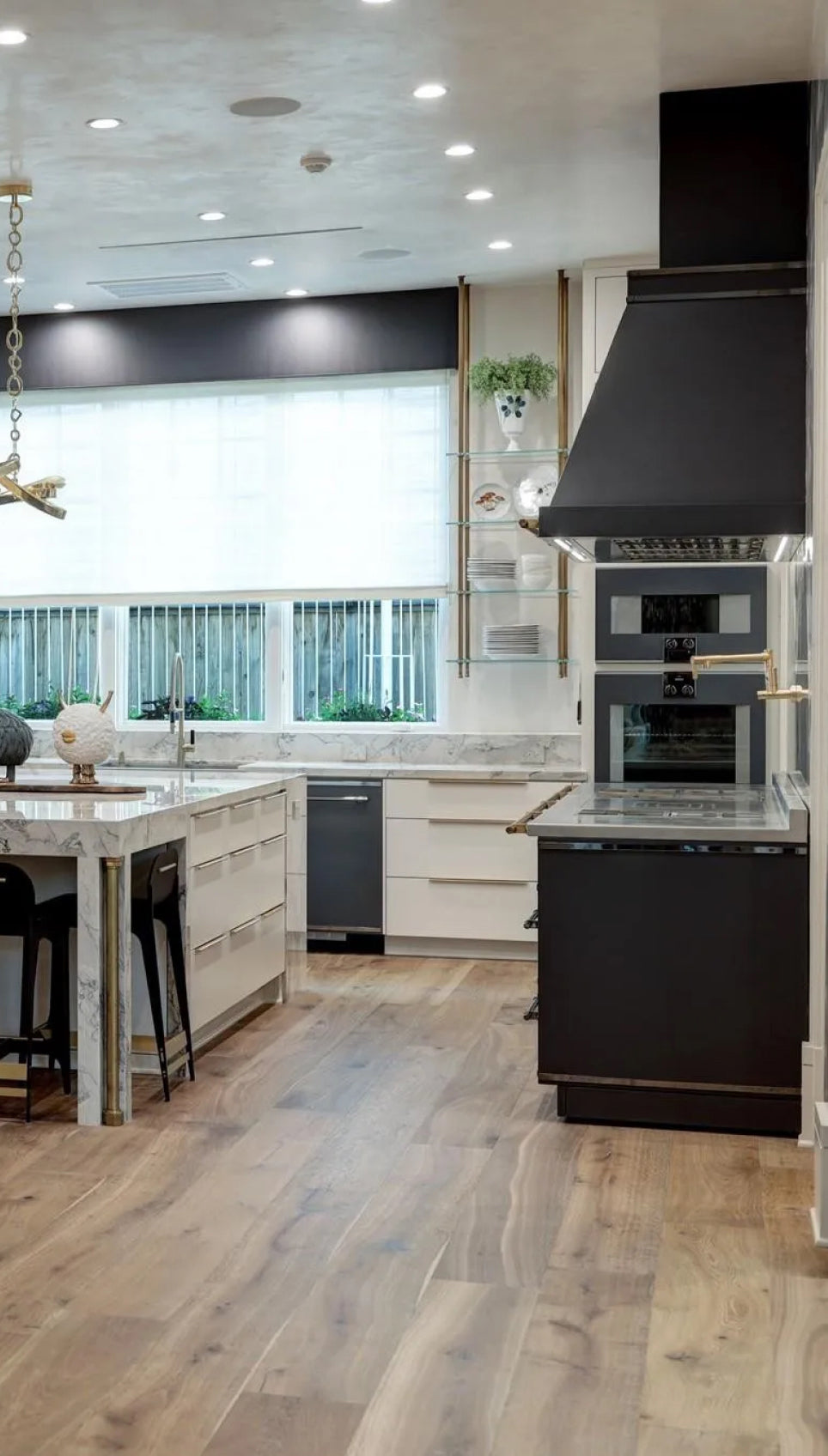 Modern L'Atelier Paris matte black kitchen featuring a professional range, matching custom ventilation hood, and integrated wall ovens, paired with a white marble waterfall island and light wood flooring.