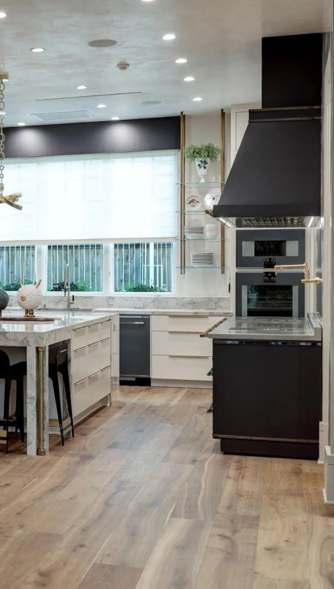 A modern and elegant kitchen with a bold black range hood and a black and white cooking range. The space features white cabinets with brass hardware, a large island with a marble countertop, and herringbone wood floors.