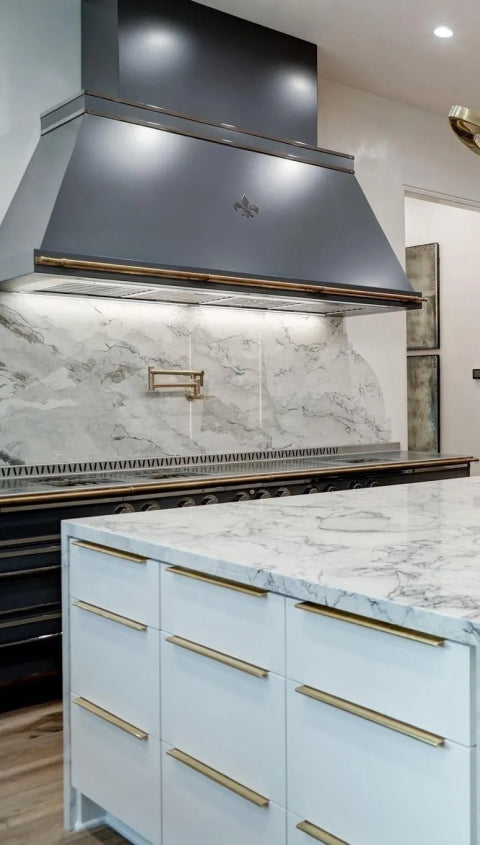 A modern kitchen featuring a large gray range hood with gold trim and a fleur-de-lis emblem, set against a striking gray-veined marble backsplash. The kitchen island in the foreground has a white marble countertop with a waterfall edge and gold hardware on the drawers.