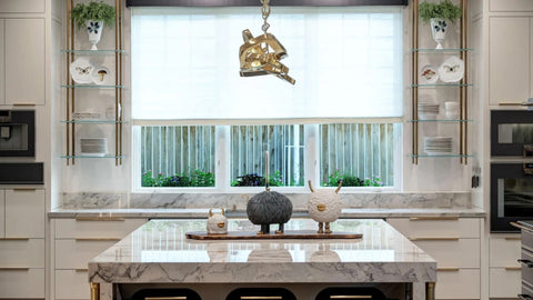 A modern and elegant kitchen featuring a large marble island with a waterfall edge and a display area for decorative items. The space has white cabinets with gold hardware, marble countertops, and glass shelves with brass frames that flank a large window.