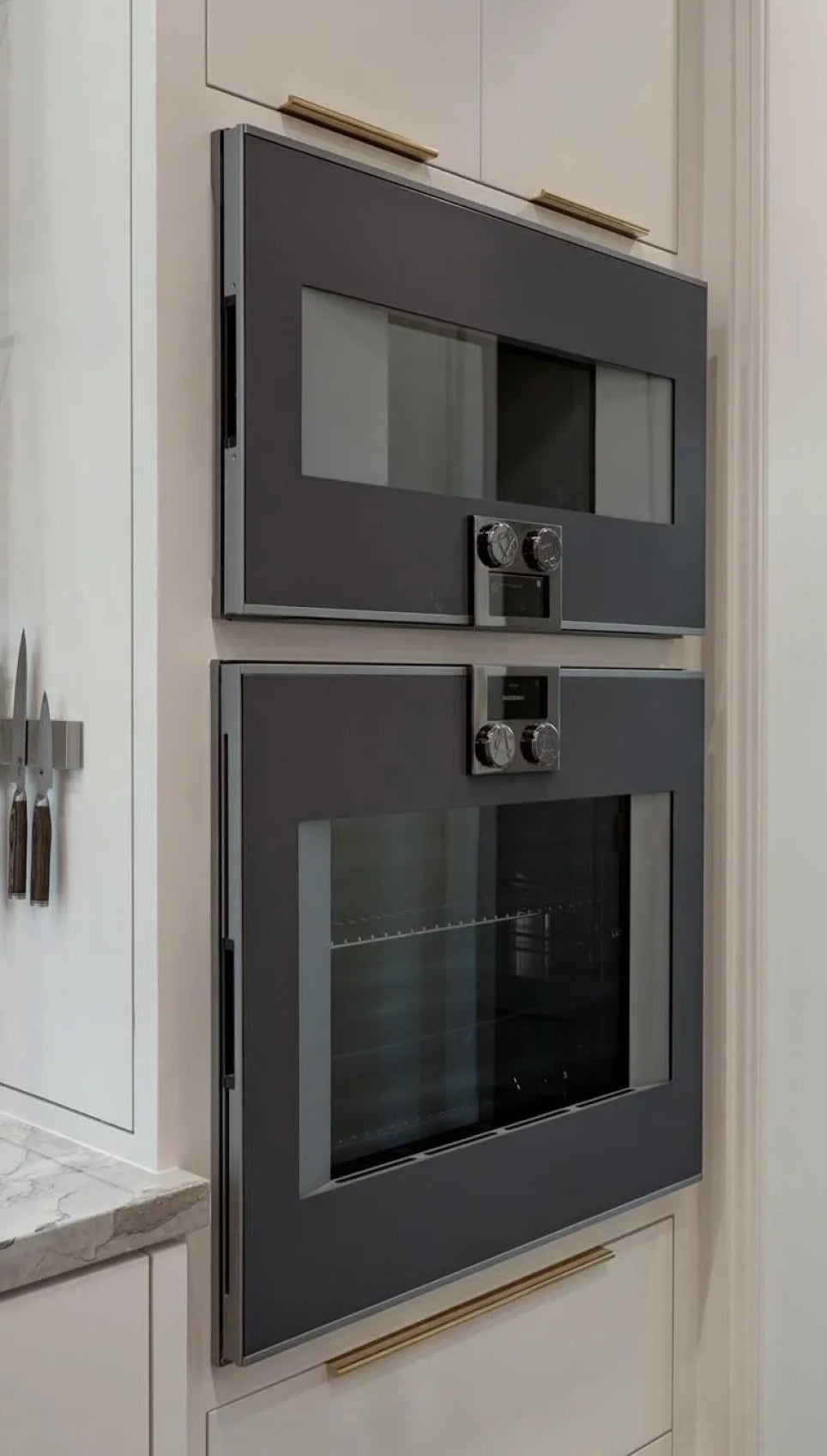 A close-up of a modern kitchen's built-in wall ovens, featuring two sleek, dark-gray ovens stacked vertically with stainless steel knobs and handles. The ovens are set into a light-colored kitchen cabinet, with a magnetic knife strip visible to the left.