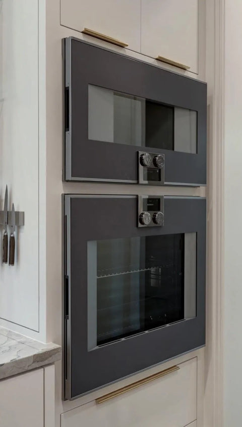 A stylish modern kitchen with a focus on appliances, showcasing a pair of stacked, dark gray built-in ovens with a clean, minimalist design and brass cabinet pulls nearby.