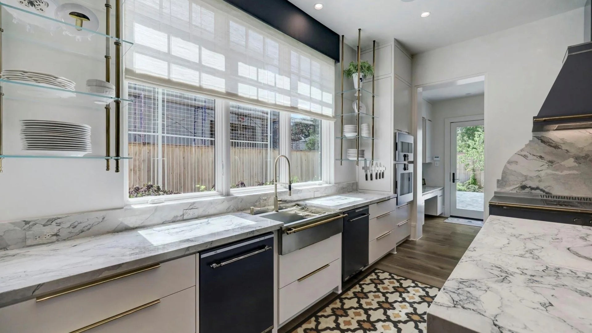 A stunning white kitchen with marble countertops and backsplash, light wood floors, and a large window above a farmhouse sink. The room is accented with brass hardware, floating glass shelves, and a patterned rug in front of the dishwasher.