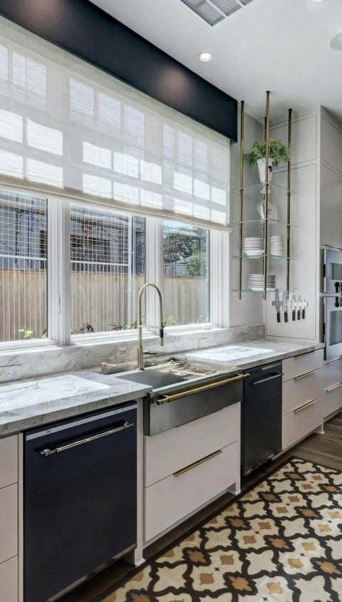 A close-up of a modern kitchen's sink area, showing a large window with white blinds, a farmhouse sink, and white marble countertops. The space is styled with a patterned floor rug, a floating glass shelf with brass accents, and a built-in knife block.
