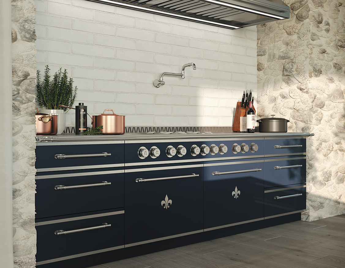 Close-up of a luxurious navy blue La Cornue range and base cabinetry with silver drawer pulls and Fleur de Lis accents, set against a wall featuring white subway tile and natural stone siding, all topped with a stainless steel range hood.