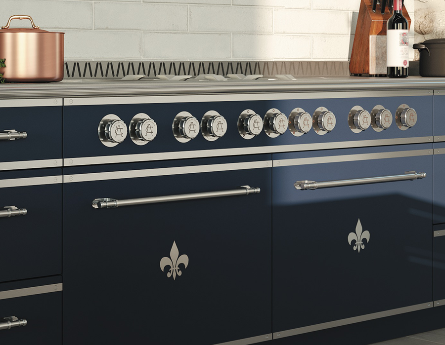 Close-up of a navy blue luxury range cooker by La Cornue or similar brand, showing the front panel with silver Fleur de Lis accents, eleven chrome control knobs, and long silver towel bar handles across the oven doors, with a copper pot visible on the counter.