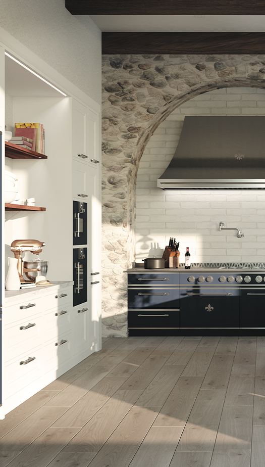 Vertical view of a luxury French kitchen with light wood flooring, showing tall white cabinetry with built-in ovens and a copper stand mixer, next to a navy blue La Cornue range and stainless steel range hood set in a dramatic stone and white brick arched alcove.