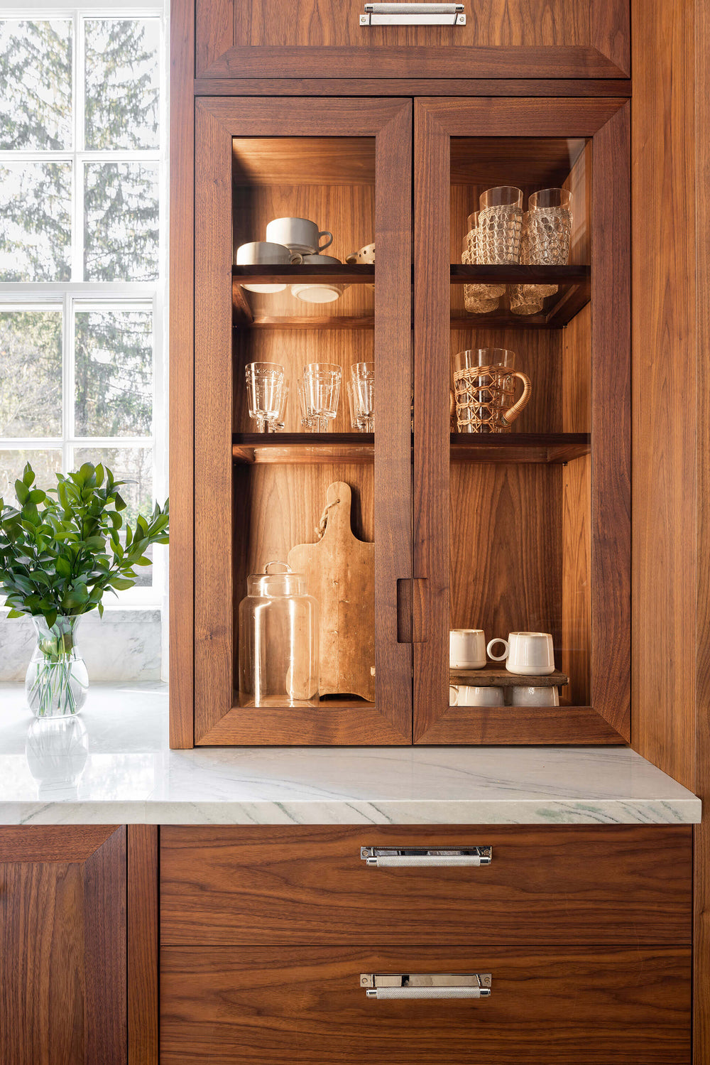 Luxury walnut wood kitchen cabinetry featuring a glass-front upper cabinet displaying glassware, mugs, and a wooden cutting board above a white marble countertop.