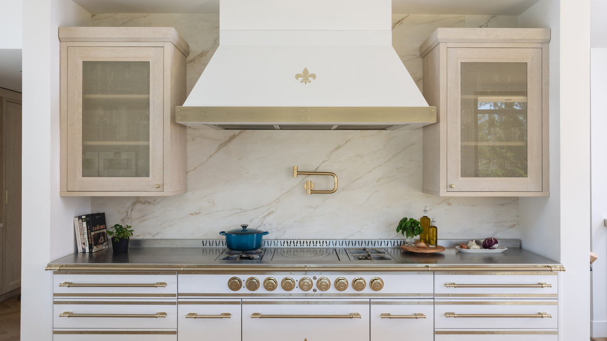 Elegant kitchen with a large white and gold range, a custom white range hood with a fleur-de-lis emblem, and a beautiful veined marble backsplash with a gold pot filler faucet.