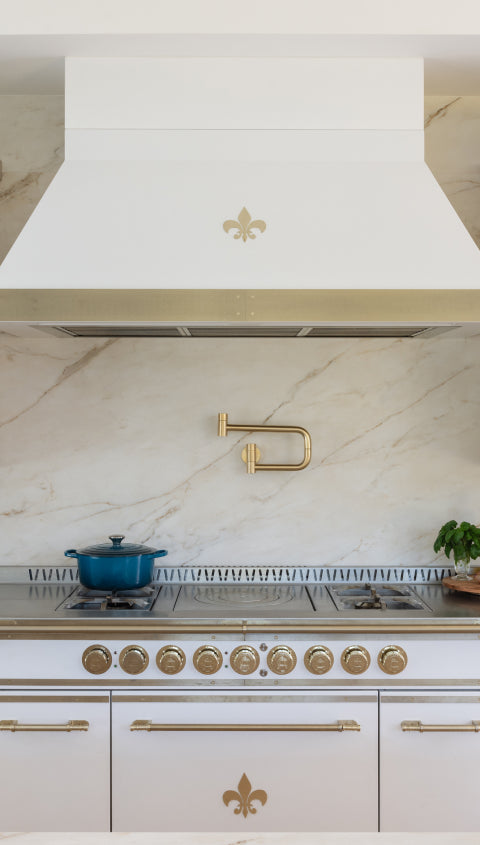 A detailed shot of a kitchen's cooking area, highlighting a white range with gold knobs and a fleur-de-lis emblem. The custom white range hood above it also features a matching gold fleur-de-lis, set against a stunning veined marble backsplash with a gold pot filler.