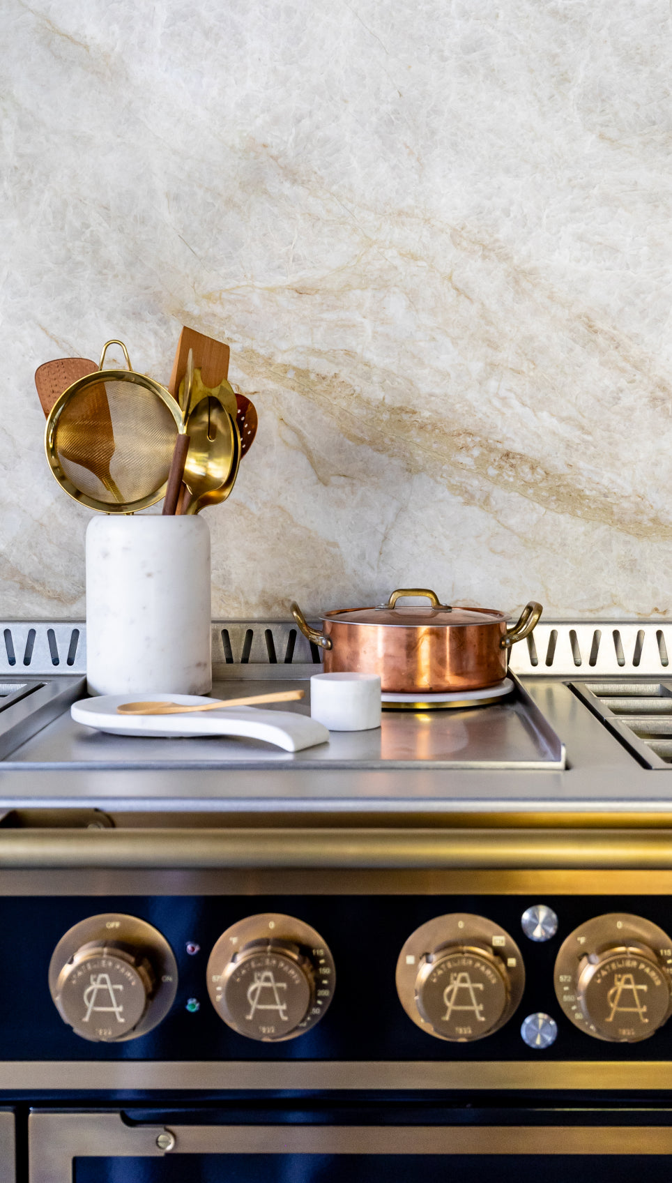 Close-up of a black L'Atelier Paris kitchen range stovetop featuring custom brass control knobs and a small copper pot