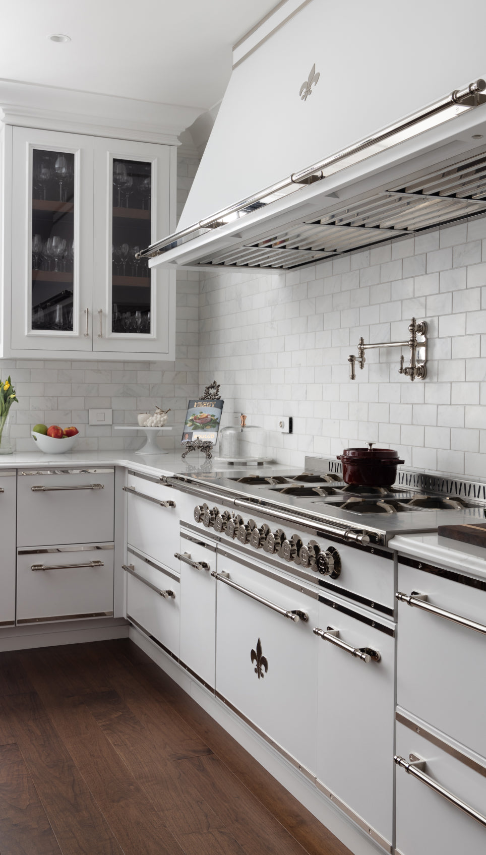 L'Atelier Paris white professional French range and matching custom ventilation hood with polished stainless steel trim and fleur-de-lis accents, integrated into white cabinetry with a marble subway tile backsplash and chrome pot filler.