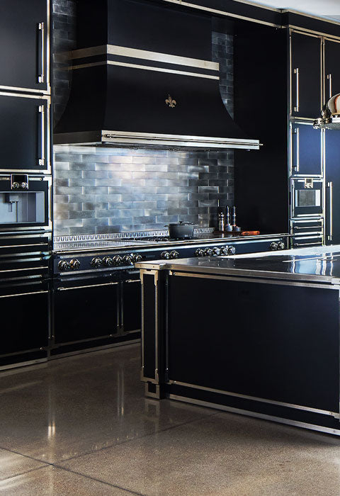 Ultra-modern luxury kitchen featuring all-black cabinetry and a large kitchen island with sleek stainless steel or chrome trim and hardware, a black La Cornue range and custom black range hood with a Fleur de Lis accent, an industrial metal subway tile backsplash, and two chrome pendant lights.