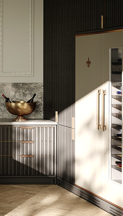 Vertical close-up of a luxury kitchen corner featuring a tall light-beige refrigerator or wine cooler with a gold Fleur de Lis emblem and long brass handles, next to dark fluted base cabinets with grey marble countertop and a decorative brass wine cooler bowl.