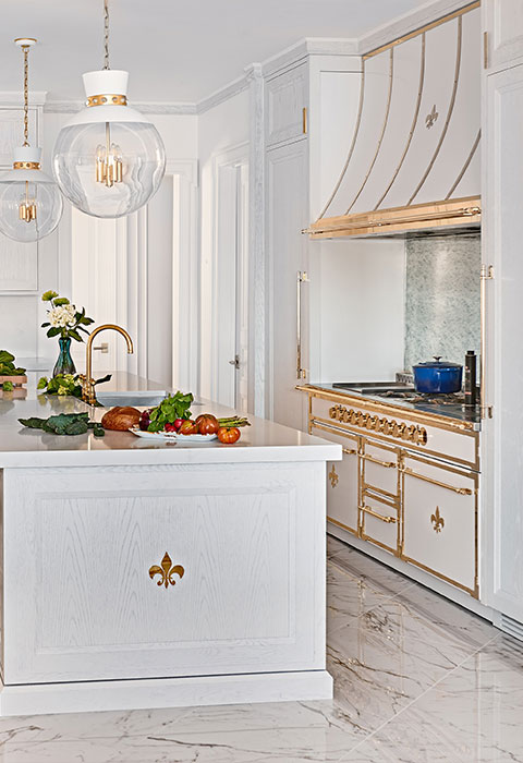 A vertical shot of a luxurious, white and gold kitchen with a French aesthetic. The focal point is a professional-style range and custom range hood, both finished in glossy white and extensively trimmed with polished gold-tone brass. A kitchen island in the foreground has a white wood-paneled base and a sink with a gold gooseneck faucet. The kitchen features marble flooring and white cabinetry with gold hardware, completing the opulent, bright design.