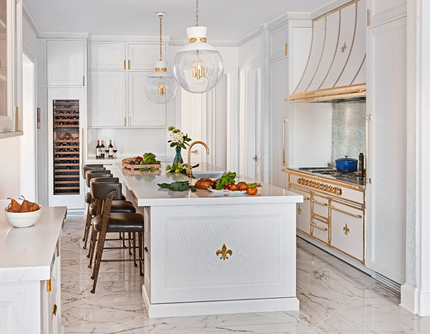 Bright white gourmet kitchen with luxury gold-trimmed French-style range, large island seating with dark stools, built-in glass-door wine refrigerator, and elegant white marble floor.