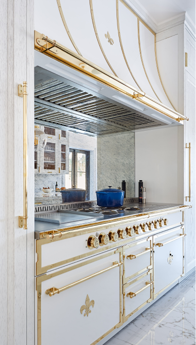 A stunning vertical close-up of a luxury French-style kitchen range and hood. The appliance features a glossy white enamel finish lavishly trimmed with polished gold-tone brass hardware, including ornate knobs, handles, and decorative frames. The oven features a Fleur-de-lis emblem on one panel. Above the range, the custom hood is also white and gold, and the backsplash is a mirrored surface reflecting the opposite side of the kitchen, which has white cabinets and marble counters.