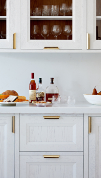 A vertical close-up of a white kitchen cabinet section featuring both glass-front upper cabinets displaying stemware and glasses, and lower drawers/cabinets. The cabinetry has a subtle wood grain texture and is accented with modern, horizontal gold-tone bar pulls. The countertop holds various items, including several liquor bottles, a white bowl of pears, and bread.