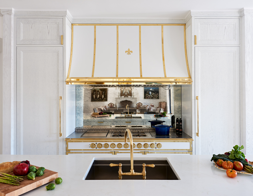White and gold luxury kitchen, symmetrical range hood, mirrored backsplash, gold faucet, white kitchen island.