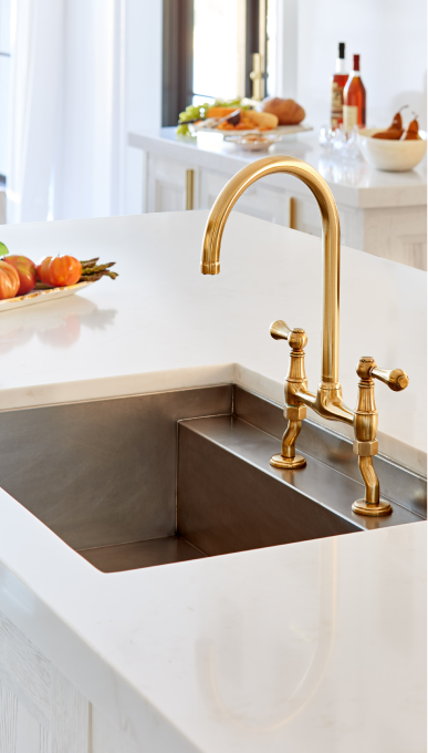 A vertical close-up of a sleek kitchen island sink with a polished gold-tone bridge faucet and cross-handle controls. The sink itself is a modern, dark metallic double basin, integrated into a clean white quartz countertop. In the blurred background, fresh vegetables and other kitchen items are visible, hinting at a bright and functional gourmet space.