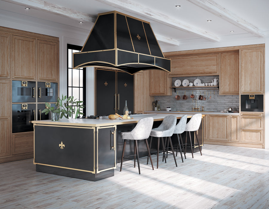 A stunning, spacious kitchen with a dramatic black and brass professional range hood as the focal point over a large kitchen island. The room features natural wood cabinetry, built-in black appliances with brass accents, a light wood floor, and a blue-gray subway tile backsplash.