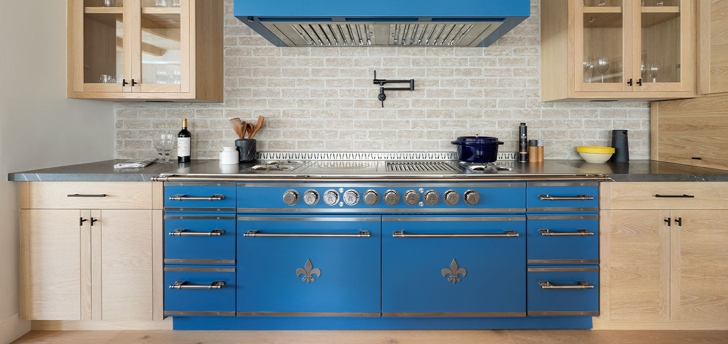 Blue kitchen cabinets with silver stove burners and handles between wooden drawers and base kitchen cabinets. Wooden hanging cabinets and blue custom kitchen hood between them