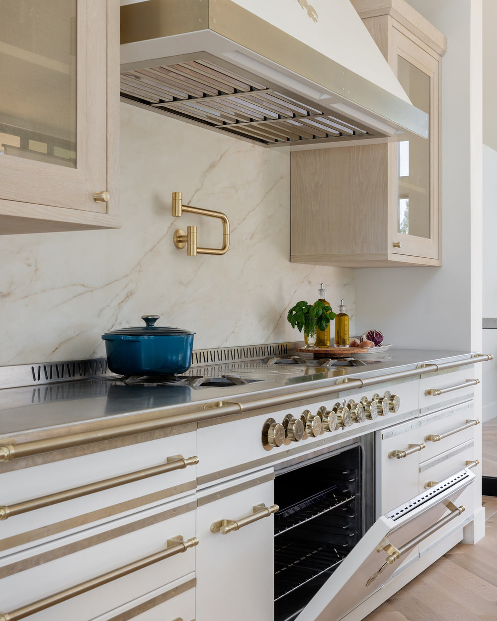 L'Atelier Paris white professional French range with an open oven door, featuring brushed brass trim, fleur-de-lis accents, and a matching gold pot filler against a marble backsplash with light wood cabinetry.