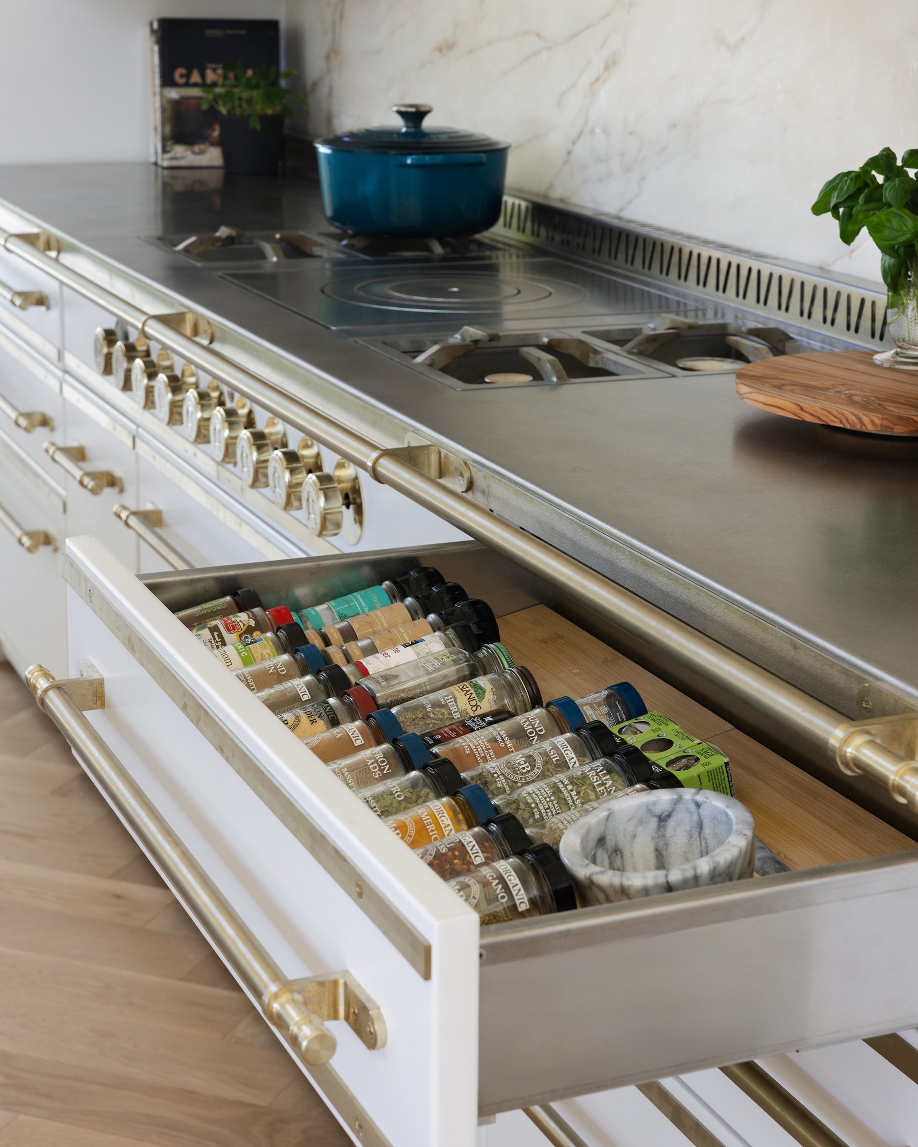 Close-up of a L'Atelier Paris white professional French range featuring an open custom spice drawer organizer filled with jars, beneath a stainless steel cooktop with brass trim.