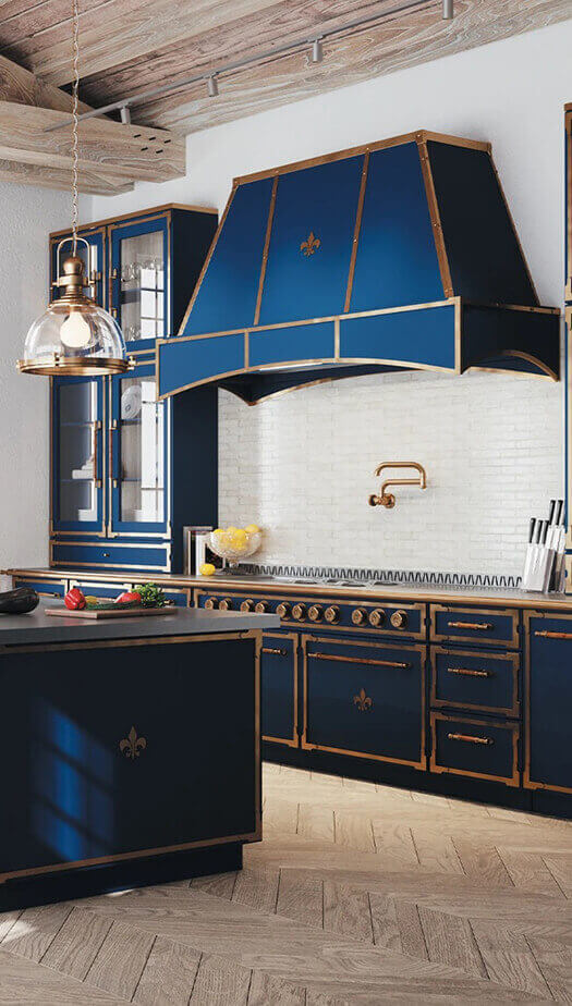 A striking, luxurious kitchen featuring deep blue cabinetry and a professional range hood accented with polished brass trim and fleur-de-lis emblems. The space includes a large kitchen island with a dark countertop and a brass faucet, a white brick backsplash with a brass pot filler, and a rustic exposed beam ceiling above a light wood floor.