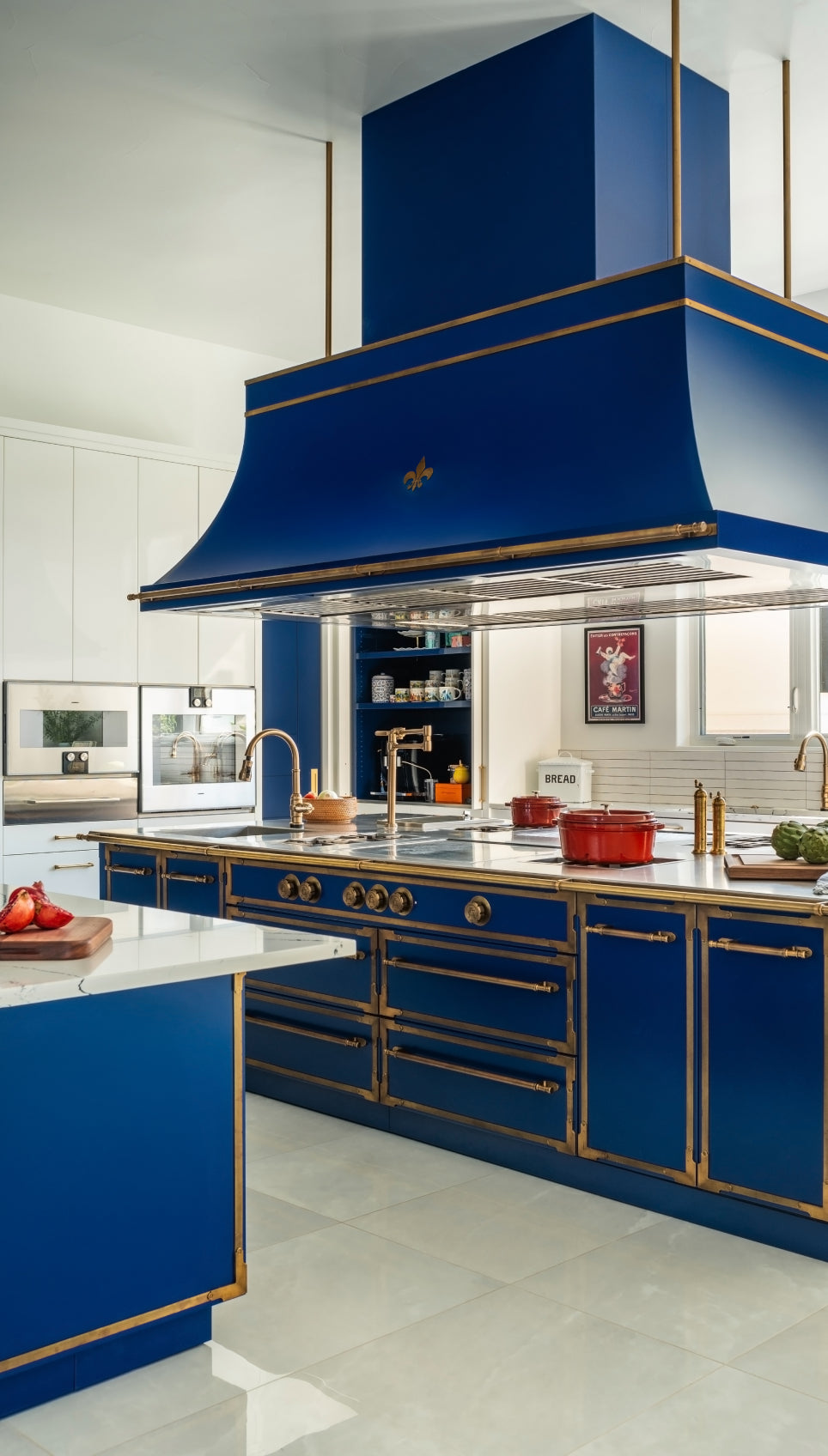 Vibrant blue custom kitchen with a large island, gold faucets, and an oversized decorative vent hood accented with brass detailing and a gold fleur-de-lis emblem.