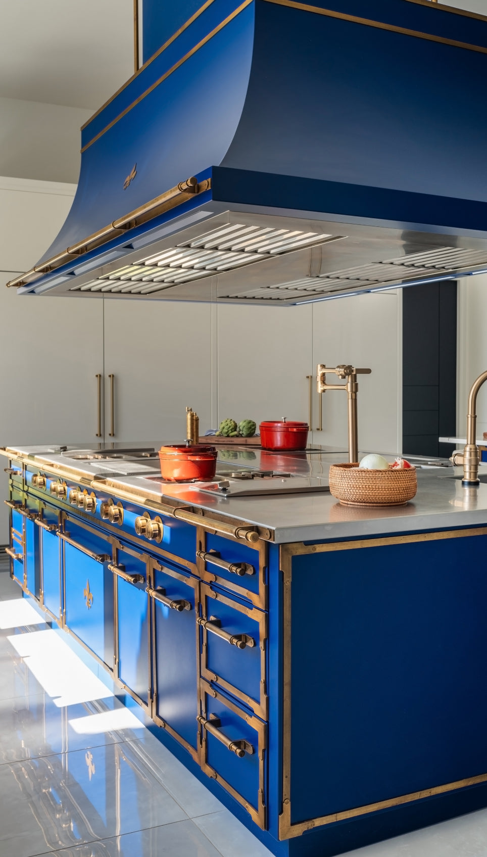 Luxury L'Atelier Paris cobalt blue kitchen island with burnished brass trim and fleur-de-lis accents, featuring an integrated stainless steel cooktop, matching professional range hood, and a gold pot filler.
