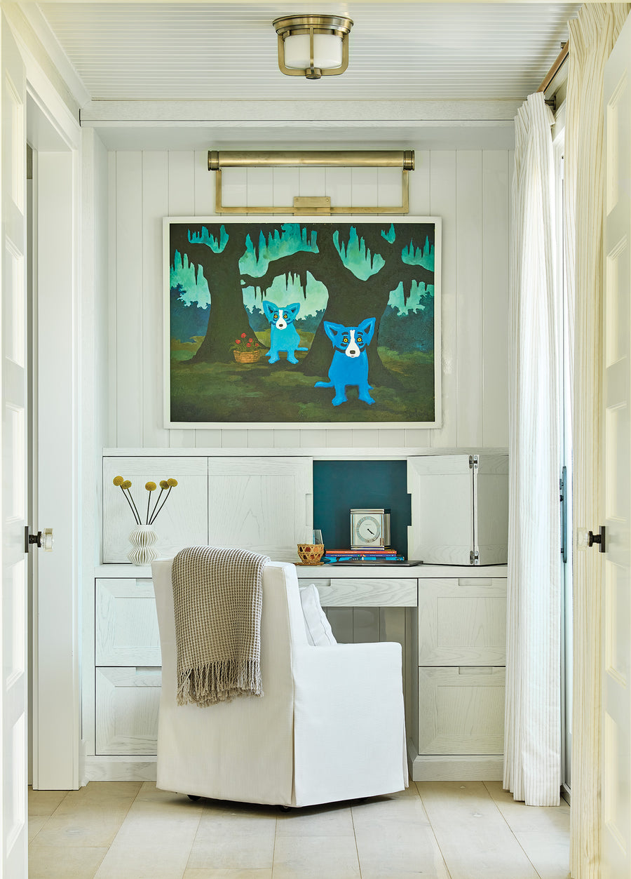 White built-in desk with brass gallery light, framed artwork, and slipcovered chair.