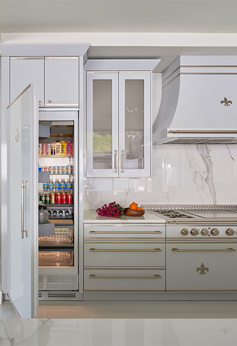 A high-end kitchen with a built-in, stainless steel refrigerator that has its door open, revealing a full stock of drinks and a glass-front freezer drawer below.