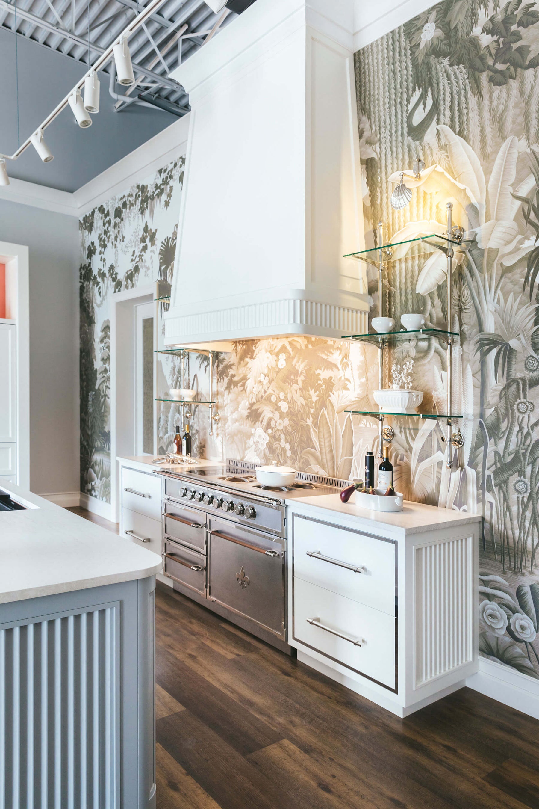 A beautiful gourmet kitchen featuring a striking monochromatic botanical wallpaper mural as the main wall covering and backsplash. The space includes a professional stainless steel range with a Fleur-de-lis emblem and a large custom white range hood. The walls are accented with glass shelving supported by polished silver-tone brackets, and the lower cabinets are a mix of white and light gray with silver pulls.