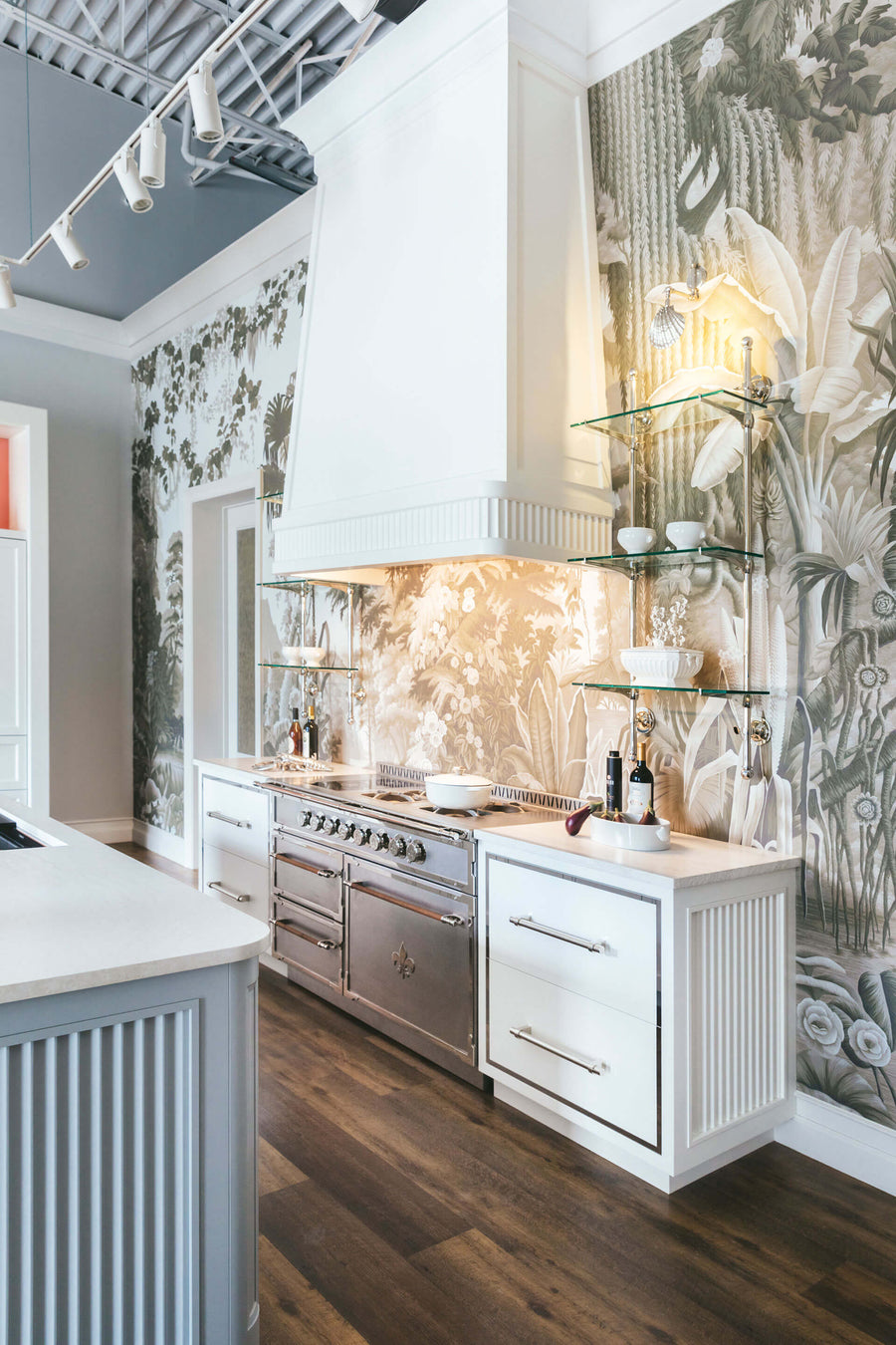 A beautiful gourmet kitchen featuring a striking monochromatic botanical wallpaper mural as the main wall covering and backsplash. The space includes a professional stainless steel range with a Fleur-de-lis emblem and a large custom white range hood. The walls are accented with glass shelving supported by polished silver-tone brackets, and the lower cabinets are a mix of white and light gray with silver pulls.