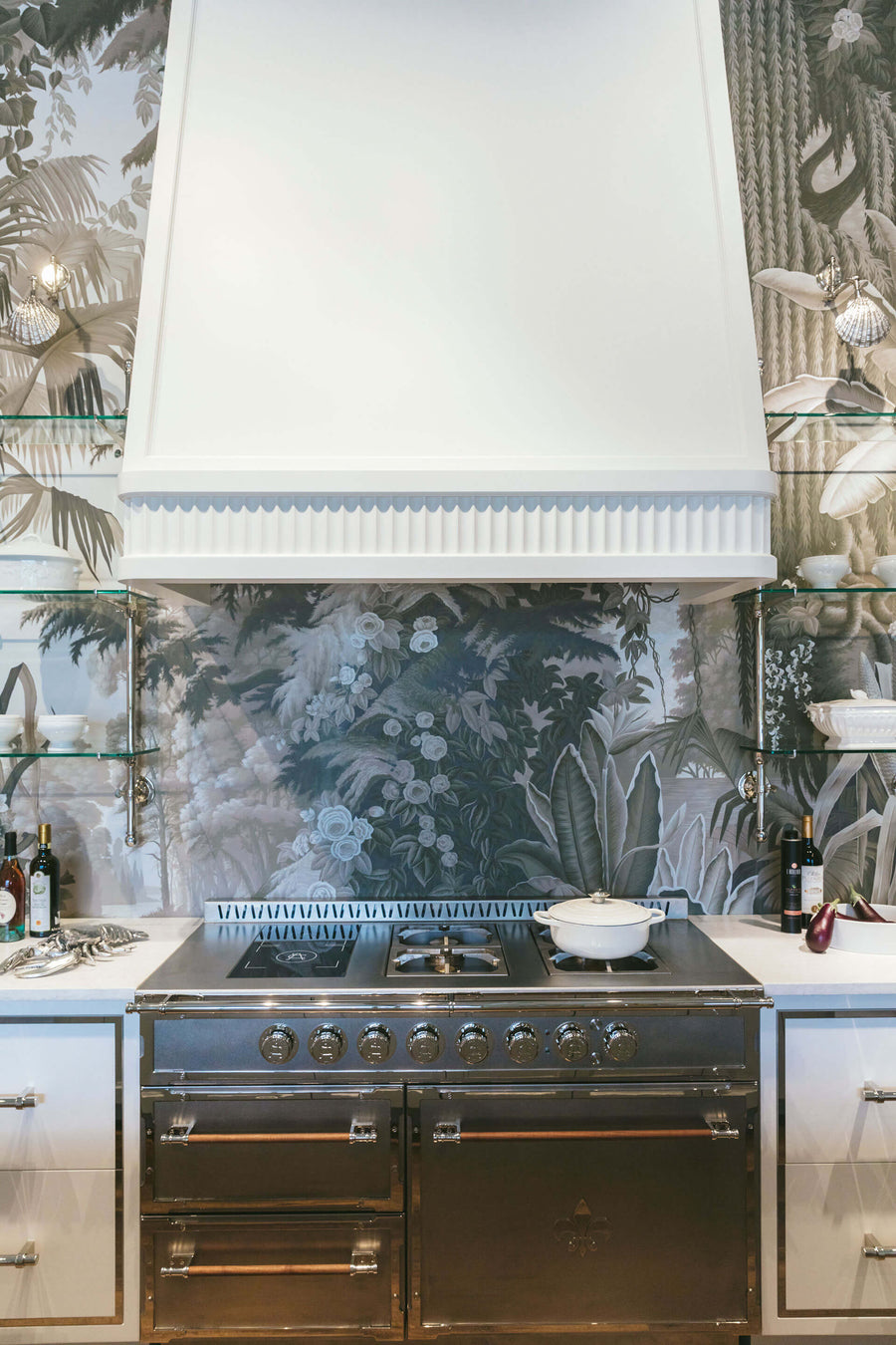 A dramatic vertical shot of a professional stainless steel range with a matte black base set against a monochromatic botanical mural wallpaper backsplash. A large custom white range hood with fluted trim is mounted above, with integrated lighting highlighting the scene. The range is flanked by glass shelving and lower cabinets, blending high-end appliance functionality with elegant, nature-inspired design.