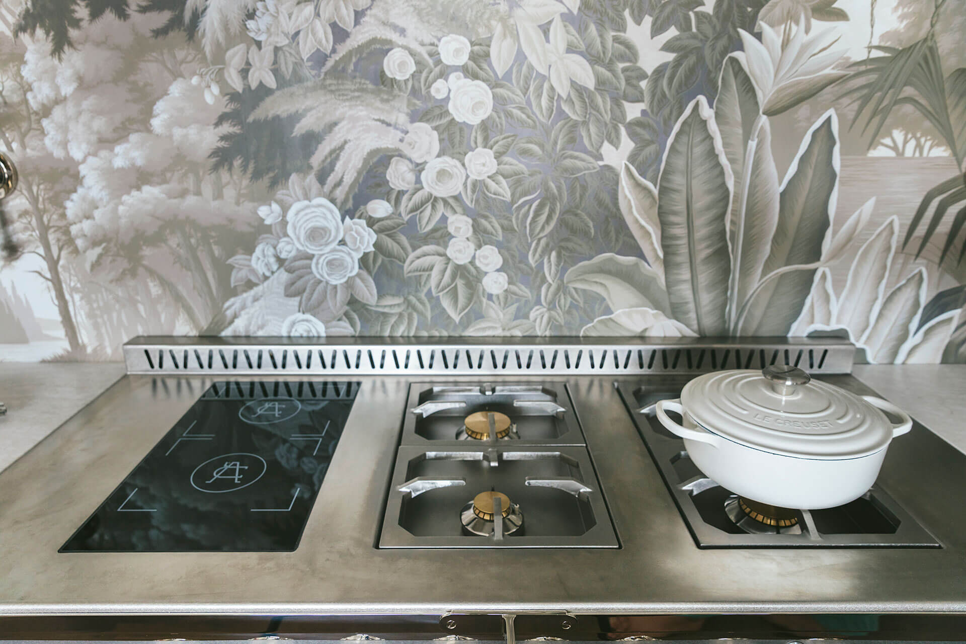 A close-up shot of a professional-grade kitchen cooktop set into a stainless steel counter. The cooktop is a hybrid design featuring a black induction zone on the left and two gas burners in the center and right. A white Le Creuset Dutch oven rests on one of the gas grates. The backdrop is a dramatic monochromatic botanical wallpaper in shades of gray and white, creating an elegant contrast with the modern appliance.