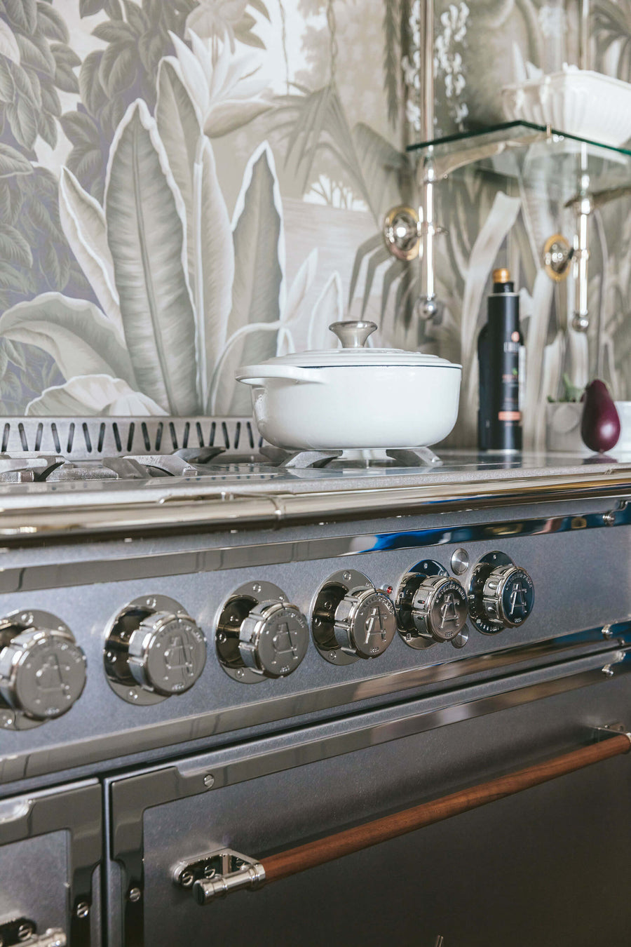 A detailed vertical close-up of a professional-grade kitchen range with a sleek stainless steel finish and large, polished chrome control knobs. A white enameled pot rests on the cooktop. The range features a dark wooden handle, and the backdrop is a striking monochromatic botanical wallpaper mural in shades of gray, with modern glass shelving visible on the right.