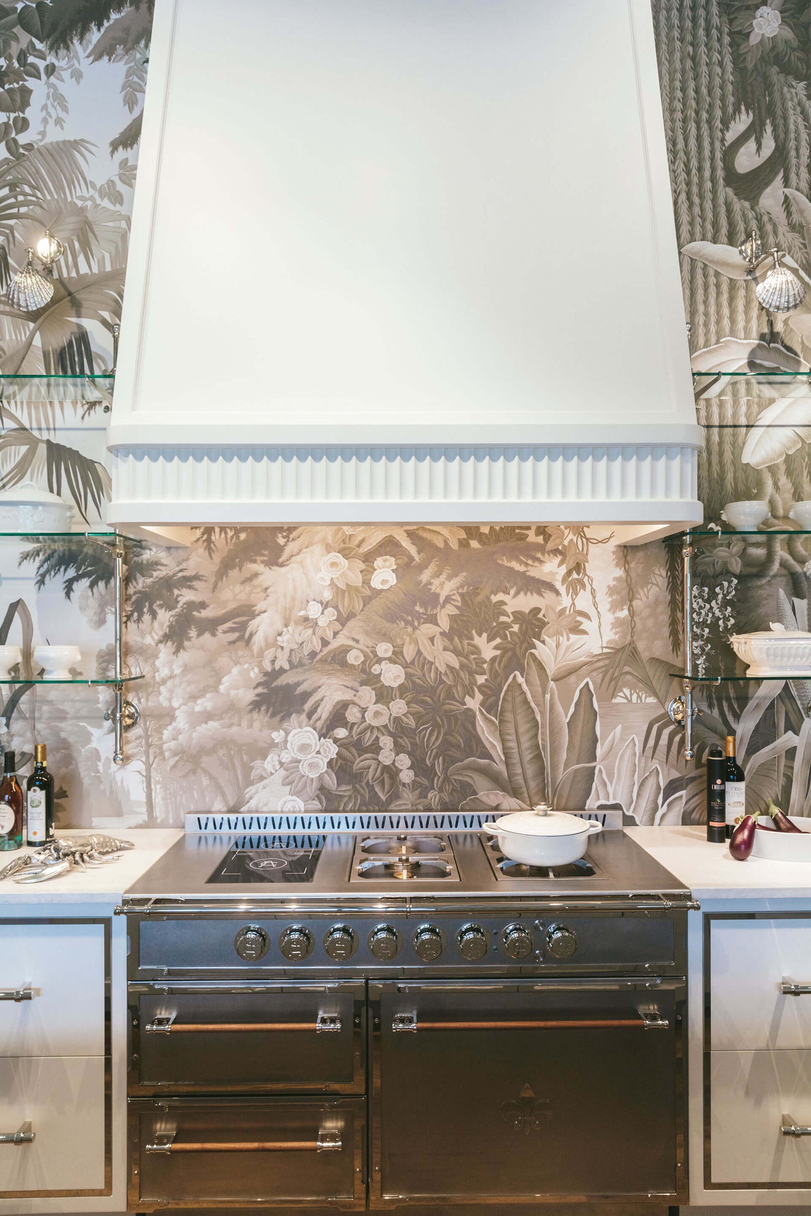 A dramatic vertical shot of a professional stainless steel range with a matte black base set against a monochromatic botanical mural wallpaper backsplash. A large custom white range hood with fluted trim is mounted above, with integrated lighting highlighting the scene. The range is flanked by glass shelving and lower cabinets, blending high-end appliance functionality with elegant, nature-inspired design.