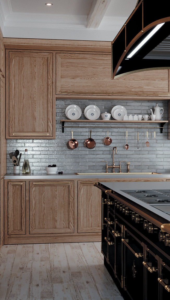 A vertical shot of a luxurious kitchen featuring natural wood cabinetry with gold hardware, a shimmering blue-gray brick-style subway tile backsplash, and a white marble-look countertop. The wall decor includes a wooden shelf with white dishes, and a rack of hanging copper pots above a brass sink faucet. A portion of a black and brass professional range is visible in the lower right.