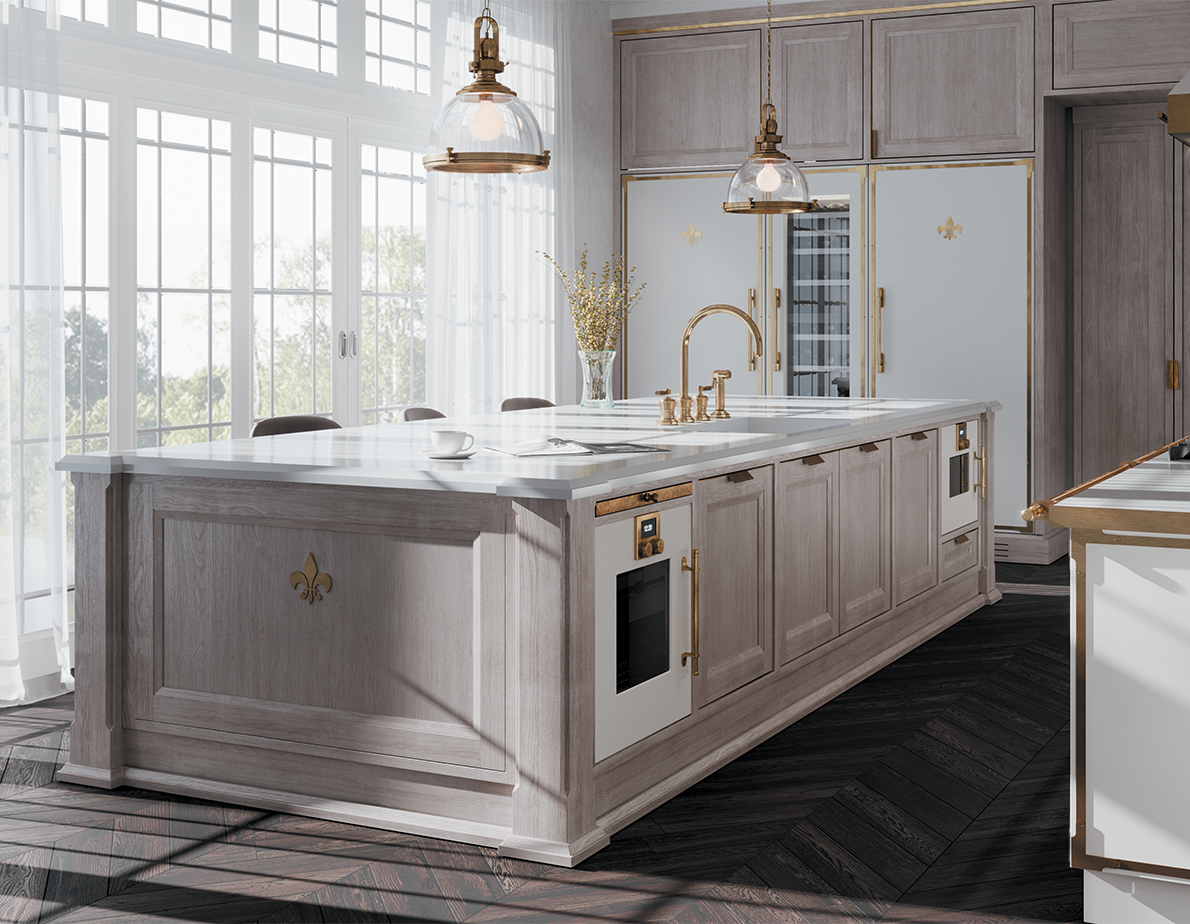 A bright, elegant kitchen centered on a large light gray wood island with a white marble countertop, gold hardware, and a fleur-de-lis emblem. The island features a built-in oven, is lit by two industrial-style pendant lights, and sits on a dark wood herringbone floor in front of large, sunlit windows with sheer white curtains.